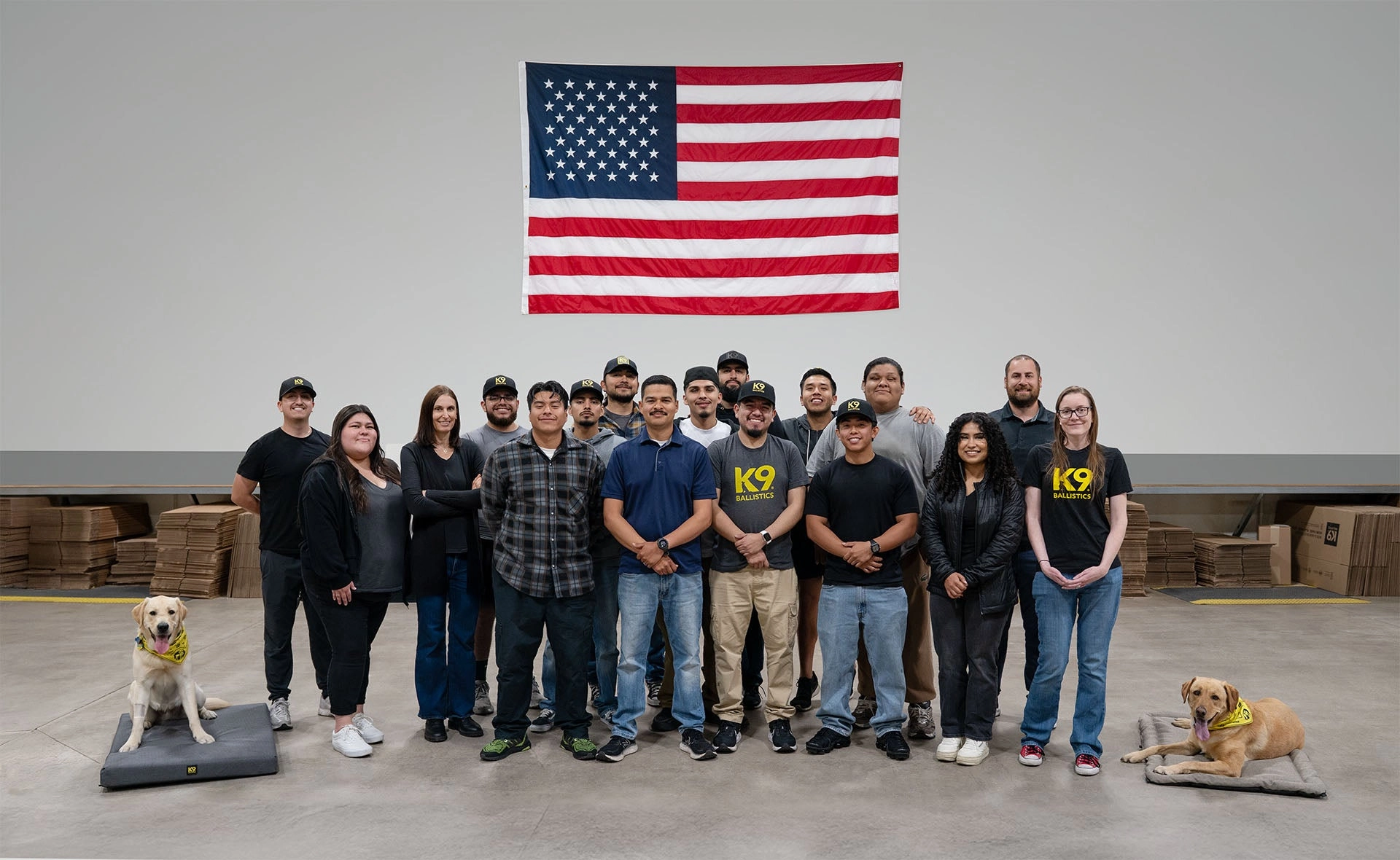 The K9 Ballistics team standing together in front of an American flag, with two dogs resting on beds in the foreground